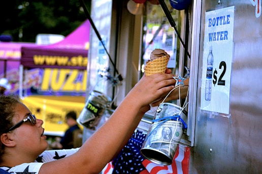 A person is reaching towards a food truck window, holding a waffle cone with ice cream. There are various decorations including stars and American flags. A sign advertises bottled water for $2, and a jar with money is visible hanging by the window.