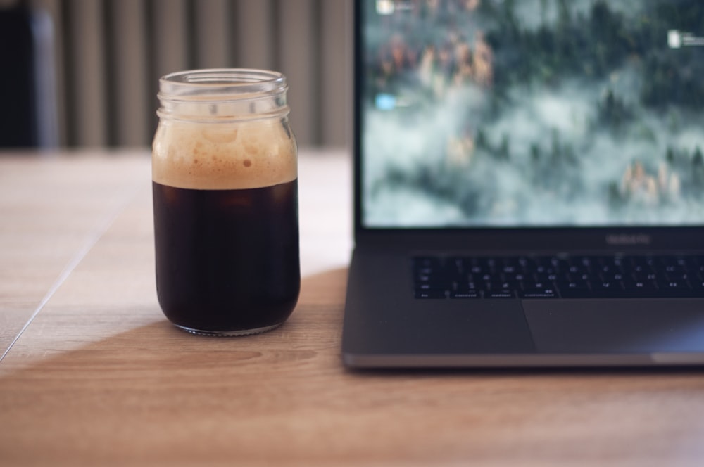clear glass mason jar filled with black liquid beside turned-on laptop