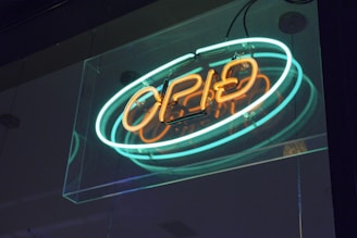 A neon sign with the word 'open' written in bright orange and blue lights is mounted on a glass surface. The sign is illuminated, casting a subtle glow in the surrounding area, creating a vibrant and inviting atmosphere.