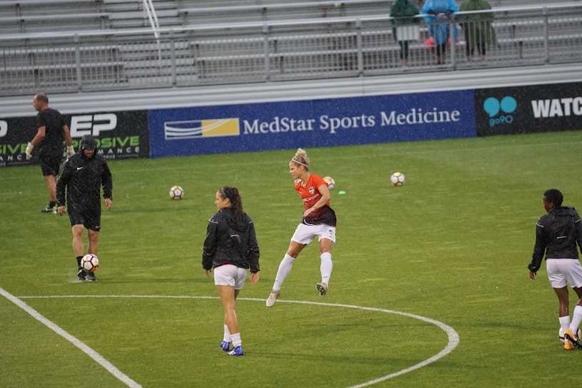 Players in sports attire are on a grassy field, engaged in soccer practice. Some are wearing black jackets and shorts, while one player in the center wears an orange jersey. Soccer balls are scattered across the field, and the stands in the background are mostly empty with a few onlookers.