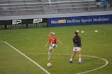 Two soccer players on a field, one in an orange jersey controlling a soccer ball with their knee, and the other in a dark jacket walking nearby. Several soccer balls and green cones are scattered on the grass. Bleachers and advertisements are visible in the background.