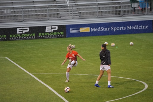 Two soccer players on a field, one in an orange jersey controlling a soccer ball with their knee, and the other in a dark jacket walking nearby. Several soccer balls and green cones are scattered on the grass. Bleachers and advertisements are visible in the background.