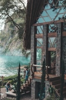 A rustic wooden and stone house situated by a tranquil lake, surrounded by lush greenery. Two people are seen engaging in outdoor activities near the steps of the house, with tall cypress trees and a rocky cliff in the background.