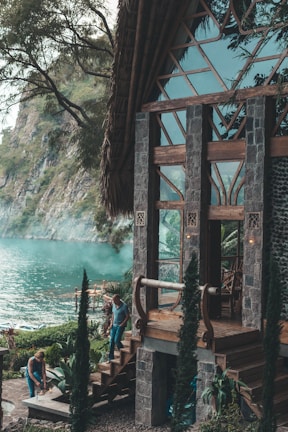 A rustic wooden and stone house situated by a tranquil lake, surrounded by lush greenery. Two people are seen engaging in outdoor activities near the steps of the house, with tall cypress trees and a rocky cliff in the background.