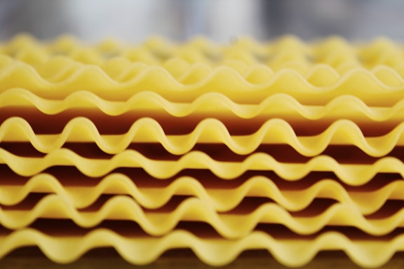 A close-up view of stacked lasagna sheets with wavy edges. The sheets are arranged in layers, creating a pattern of curves and shadows.
