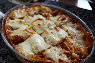 A freshly baked dish of lasagna is presented in a glass baking dish. The top is golden brown with melted cheese and crispy edges, indicating it has just come out of the oven. The layers of pasta and filling are visibly rich and inviting.