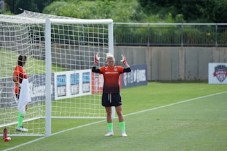 Two soccer players in orange jerseys are positioned near a goalpost on a soccer field. One player stands inside the goal, seemingly holding a towel, while the other stands outside the goal with their arms raised. The grass appears green and freshly cut, surrounded by advertising boards and a concrete wall.