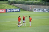 Three female soccer players in orange and black jerseys are standing on a lush green soccer field, engaged in what appears to be a discussion or planning session. Several soccer balls are scattered around them. In the background, there is a banner with social media icons and handles, located on the fence surrounding the field.