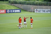 Three female soccer players in orange and black jerseys are standing on a lush green soccer field, engaged in what appears to be a discussion or planning session. Several soccer balls are scattered around them. In the background, there is a banner with social media icons and handles, located on the fence surrounding the field.