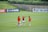 Three female soccer players in orange and black jerseys are standing on a lush green soccer field, engaged in what appears to be a discussion or planning session. Several soccer balls are scattered around them. In the background, there is a banner with social media icons and handles, located on the fence surrounding the field.