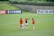 Three female soccer players in orange and black jerseys are standing on a lush green soccer field, engaged in what appears to be a discussion or planning session. Several soccer balls are scattered around them. In the background, there is a banner with social media icons and handles, located on the fence surrounding the field.
