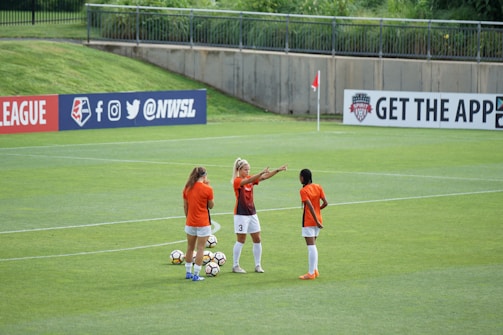 Three female soccer players in orange and black jerseys are standing on a lush green soccer field, engaged in what appears to be a discussion or planning session. Several soccer balls are scattered around them. In the background, there is a banner with social media icons and handles, located on the fence surrounding the field.