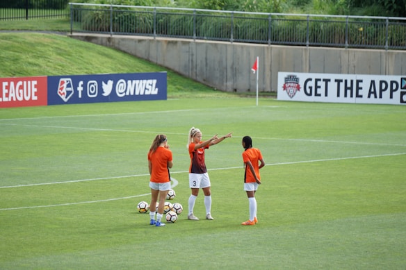 Three female soccer players in orange and black jerseys are standing on a lush green soccer field, engaged in what appears to be a discussion or planning session. Several soccer balls are scattered around them. In the background, there is a banner with social media icons and handles, located on the fence surrounding the field.