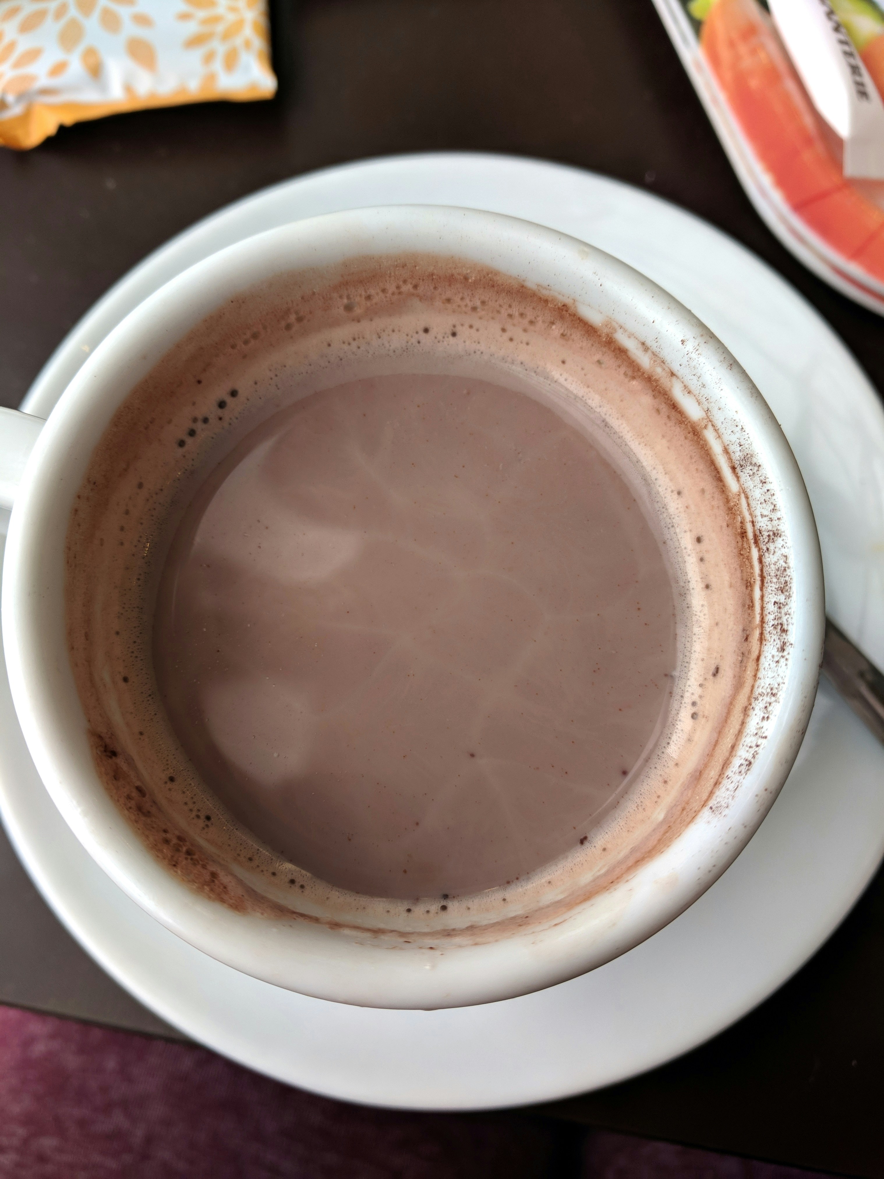 A close-up view of a cup filled with frothy hot chocolate, set on a white saucer. The surface shows slight bubbles and swirls. In the background, part of a colorful plate and a decorative napkin are partially visible.