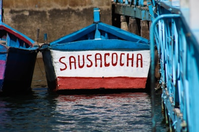 A wooden boat floating on water with the name 'Sausacocha' painted on its front in red letters. It is surrounded by blue railings and adjacent boats with similar blue and red colors. The background consists of a stone or concrete wall.