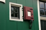 A section of a green wall with a black and white window and a red latch door. The window has a grid pattern made up of small panes, bordered with white trim. The red latch door has a square opening and is secured by black hardware. There is also a window to the right with a similar grid pattern.