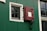 A section of a green wall with a black and white window and a red latch door. The window has a grid pattern made up of small panes, bordered with white trim. The red latch door has a square opening and is secured by black hardware. There is also a window to the right with a similar grid pattern.