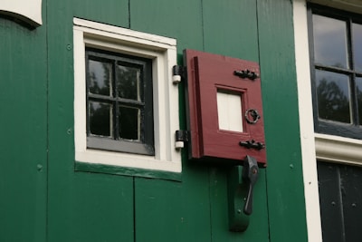 A section of a green wall with a black and white window and a red latch door. The window has a grid pattern made up of small panes, bordered with white trim. The red latch door has a square opening and is secured by black hardware. There is also a window to the right with a similar grid pattern.