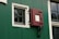 A section of a green wall with a black and white window and a red latch door. The window has a grid pattern made up of small panes, bordered with white trim. The red latch door has a square opening and is secured by black hardware. There is also a window to the right with a similar grid pattern.