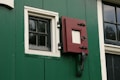 A section of a green wall with a black and white window and a red latch door. The window has a grid pattern made up of small panes, bordered with white trim. The red latch door has a square opening and is secured by black hardware. There is also a window to the right with a similar grid pattern.