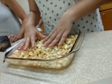 Hands are mixing chopped, seasoned apples in a clear baking dish on a speckled counter. People are standing around, partially visible, suggesting a group cooking activity.