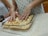 Hands are mixing chopped, seasoned apples in a clear baking dish on a speckled counter. People are standing around, partially visible, suggesting a group cooking activity.