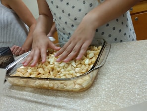 Hands are mixing chopped, seasoned apples in a clear baking dish on a speckled counter. People are standing around, partially visible, suggesting a group cooking activity.