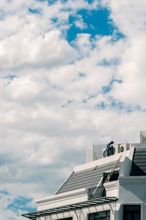 A building roof is visible under a vast sky with numerous fluffy white clouds. The structure has a modern design, with metal details and a dark, tiled roof. A person is standing on the rooftop next to some air conditioning units.