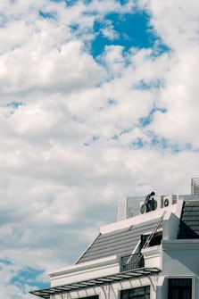 A building roof is visible under a vast sky with numerous fluffy white clouds. The structure has a modern design, with metal details and a dark, tiled roof. A person is standing on the rooftop next to some air conditioning units.