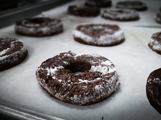 Several chocolate donuts with a dusting of powdered sugar are resting on a baking sheet lined with parchment paper. The donuts are arranged in rows and appear to be freshly baked.