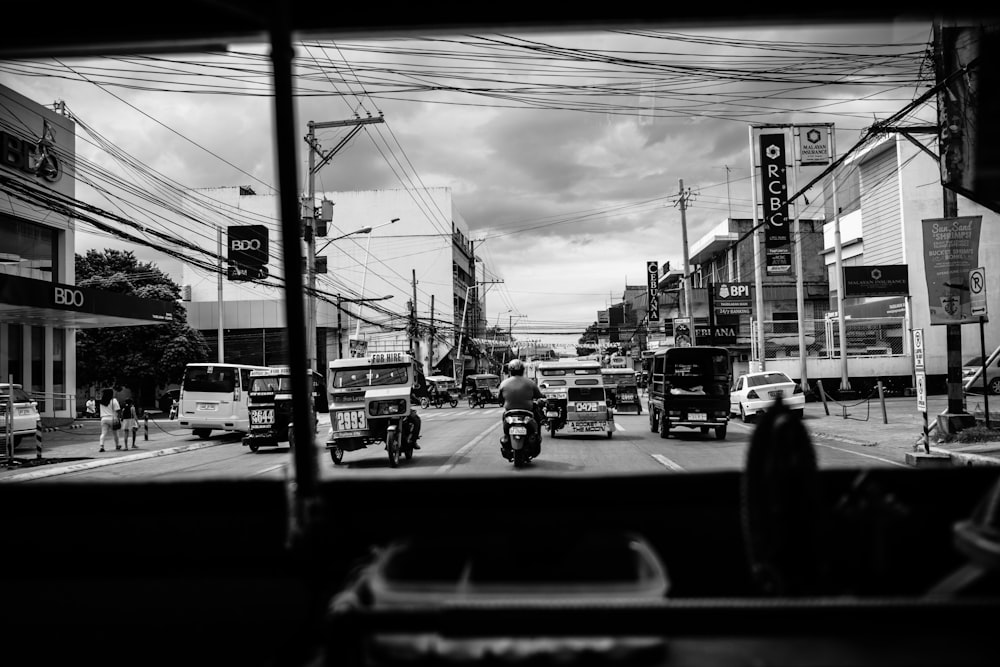 grayscale photo of man riding on motorcycle in road near buildings