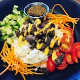 A black bowl contains a colorful dish featuring white rice topped with grilled beef drizzled with a yellow sauce. Fresh cucumber pieces, shredded carrots, sliced cherry tomatoes, and a small metal cup of dipping sauce with sesame seeds encircle the central ingredients.