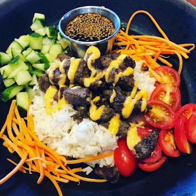 A black bowl contains a colorful dish featuring white rice topped with grilled beef drizzled with a yellow sauce. Fresh cucumber pieces, shredded carrots, sliced cherry tomatoes, and a small metal cup of dipping sauce with sesame seeds encircle the central ingredients.