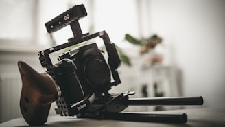 A professional camera rig with a digital camera mounted on it. The setup includes a handle, supports, and a wooden handgrip for stabilization. Background shows an out-of-focus room interior with light coming through a window.