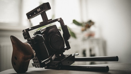 A professional camera rig with a digital camera mounted on it. The setup includes a handle, supports, and a wooden handgrip for stabilization. Background shows an out-of-focus room interior with light coming through a window.