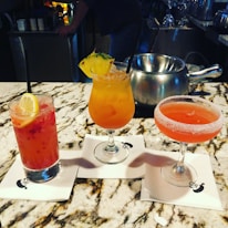 Three cocktails are arranged on a marble countertop. The drink on the left is pink with a lemon slice, the center drink is orange with a pineapple garnish, and the drink on the right is a red cocktail in a sugar-rimmed glass. Each drink is set on a napkin with a logo.