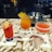Three cocktails are arranged on a marble countertop. The drink on the left is pink with a lemon slice, the center drink is orange with a pineapple garnish, and the drink on the right is a red cocktail in a sugar-rimmed glass. Each drink is set on a napkin with a logo.