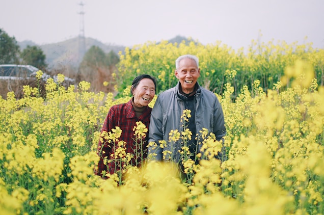 An elderly couple stands surrounded by a field of vibrant yellow flowers, smiling joyfully. The background includes blurred foliage and distant hills under an overcast sky, creating a serene and cheerful atmosphere.