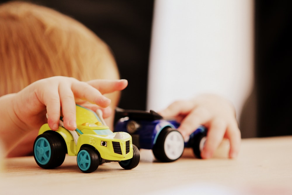 criança brincando com dois brinquedos de plástico de carro de cores variadas na mesa de madeira marrom