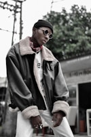 A person is wearing stylish round sunglasses, a black beanie, and a large dark leather jacket with a fur collar and cuffs. The person also sports a patterned scarf and multiple rings on their fingers. The background features green foliage and some structural elements, like a pole and a partial view of a building or outdoor furniture.