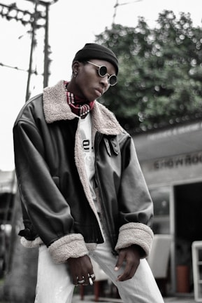 A person is wearing stylish round sunglasses, a black beanie, and a large dark leather jacket with a fur collar and cuffs. The person also sports a patterned scarf and multiple rings on their fingers. The background features green foliage and some structural elements, like a pole and a partial view of a building or outdoor furniture.