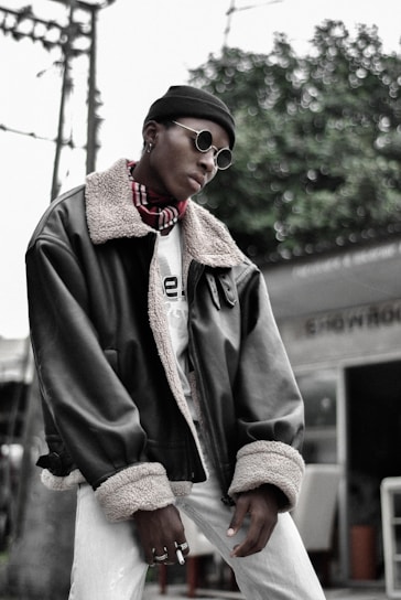 A person is wearing stylish round sunglasses, a black beanie, and a large dark leather jacket with a fur collar and cuffs. The person also sports a patterned scarf and multiple rings on their fingers. The background features green foliage and some structural elements, like a pole and a partial view of a building or outdoor furniture.
