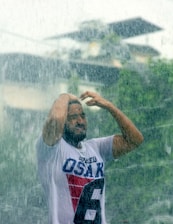 A person stands under heavy rainfall, wearing a white t-shirt with bold letters and numbers. Their hands are raised, possibly adjusting their hair or shielding their face from the rain. The rain appears intense, and the background is blurred, suggesting an outdoor setting with what looks like foliage and structures.