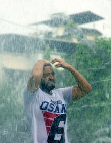 A person stands under heavy rainfall, wearing a white t-shirt with bold letters and numbers. Their hands are raised, possibly adjusting their hair or shielding their face from the rain. The rain appears intense, and the background is blurred, suggesting an outdoor setting with what looks like foliage and structures.