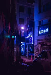 A dimly lit urban alleyway illuminated by neon lights. The scene features graffiti-covered walls, a parked car, and bright blue and purple hues. There are signs with illuminated neon lettering and reflections visible in the glass windows.