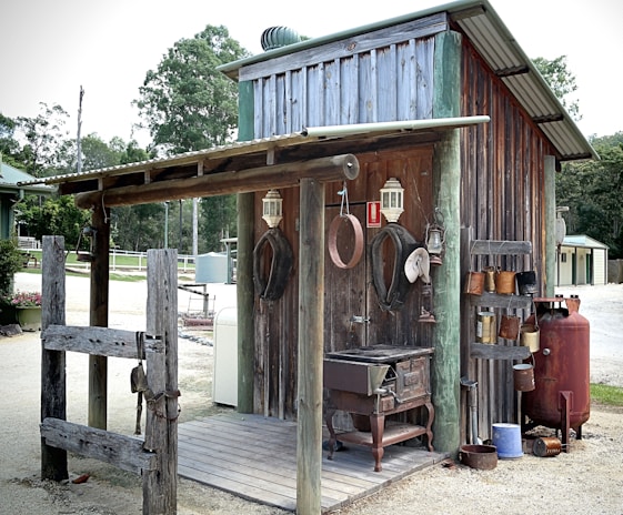 A rustic, weathered outdoor structure with a corrugated metal roof, wooden walls, and an open porch. Various vintage and rustic items are displayed, including lanterns, horseshoes, hats, and a collection of pots. An old stove sits on the porch, and a large red propane tank is positioned nearby. The setting is rural, with surrounding trees and dirt paths.