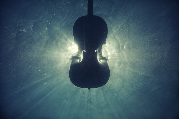 Silhouette of a cello submerged underwater, with light streaming around it creating an ethereal glow. The cello's contours are emphasized by the contrast between its dark form and the swirling lit water.