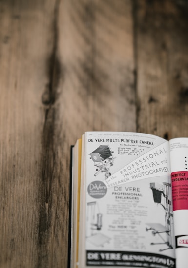 A vintage magazine or book page on a wooden surface, featuring an advertisement for a multi-purpose camera with images of camera equipment and text describing its industrial and professional use.