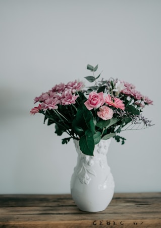 A bouquet of pink and purple flowers arranged elegantly in a white ceramic vase with embossed leaf designs. The vase is placed on a wooden table against a plain light gray background, creating a minimalistic and serene composition. The flowers appear fresh and vibrant, with greenery interspersed to enhance the arrangement.