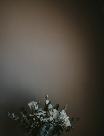 A bouquet of white flowers with green leaves is positioned against a muted, dark background. The arrangement appears delicate and elegant, with a focus on simplicity and natural elements.